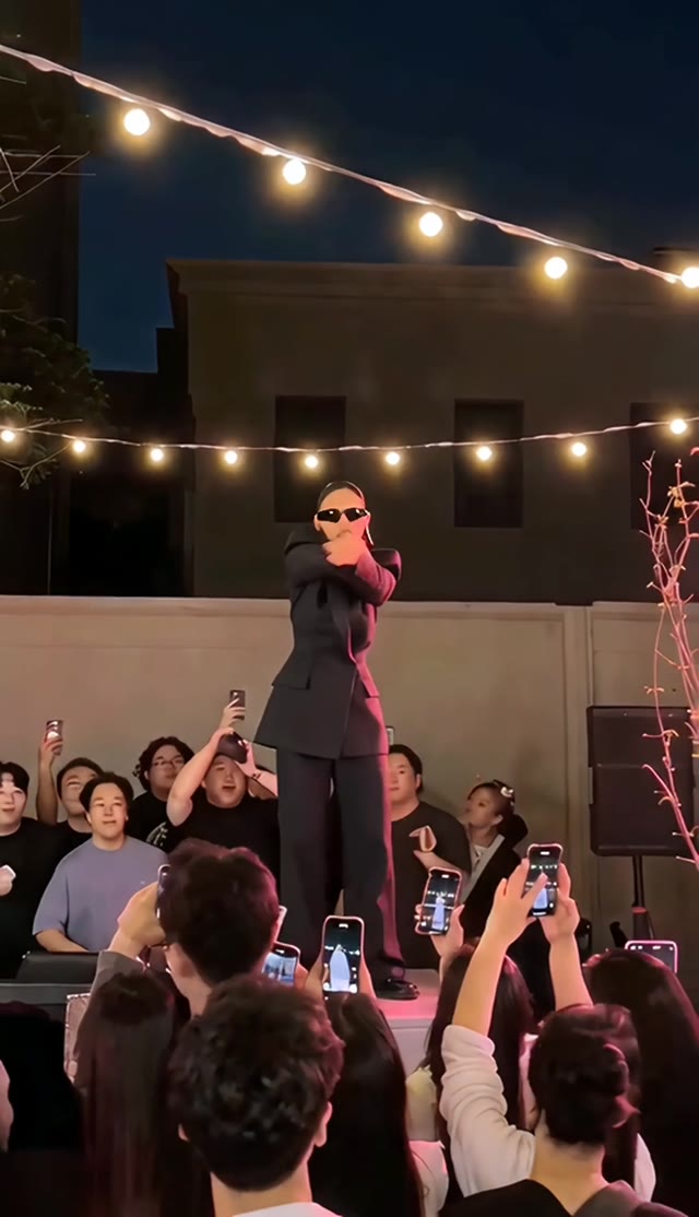 A dancer in a dark suit crosses their arms on a stage surrounded by spectators.